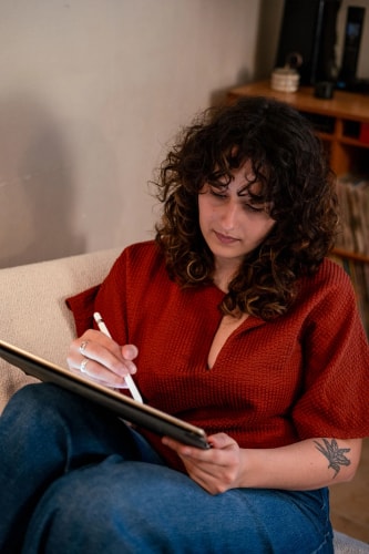 Femme aux cheveux bouclés, assise sur un canapé, utilisant une tablette graphique pour dessiner.