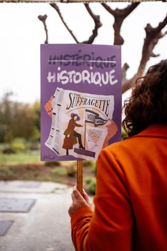 Une personne tenant une pancarte avec le texte 'Hystérique Historique' et une illustration d'une suffragette, avec des détails sur le vote.