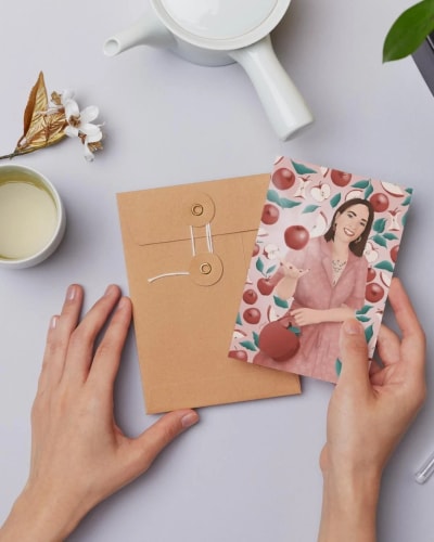 Une main tenant une carte illustrée avec une femme souriante entourée de fruits, posée à côté d'une enveloppe marron et d'une tasse de thé sur une table grise.