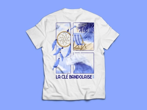 Un t-shirt blanc avec un design graphique représentant des nuages, une chaise longue, un attrape-rêves et des oiseaux. Le texte 'LA CLÉ BANDOLAISE 2024' est inscrit en bas.