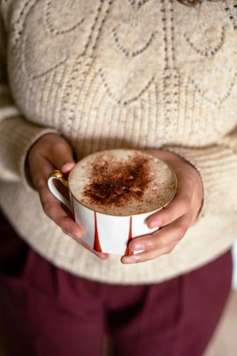 Une personne tenant une tasse de café recouverte de cacao, portant un pull beige avec un motif en tricot.
