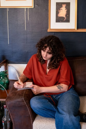 Une personne assise sur un canapé en cuir, écrivant dans un carnet avec un crayon, portant un haut rouge et un jean. Elle a les cheveux bouclés et fait face à un mur bleu foncé avec des œuvres d'art accrochées.