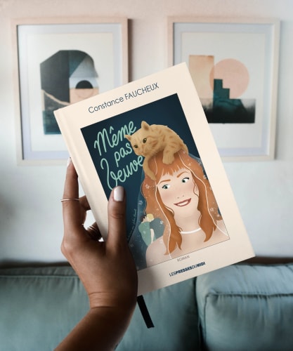 Une main tenant un livre intitulé 'Même pas Jeanne' de Constance Fauchéux, avec une couverture illustrée représentant une femme avec de longs cheveux bouclés et un chat sur sa tête, dans un intérieur moderne.