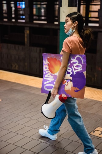Jeune femme marchant dans une station de métro, portant un mégaphone dans une main et une grande affiche colorée dans l'autre, avec un masque sur le visage.