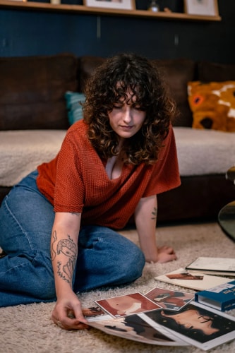 Une personne avec des cheveux bouclés et un tatouage, assise par terre sur un tapis, examinant des illustrations sur des feuilles étalées autour d'elle. Elle porte un pull rouge et un pantalon en denim.