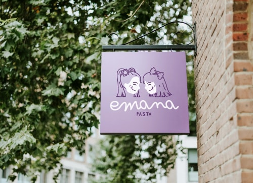 Panneau avec le texte 'emana pasta' et deux visages de filles dessinés, suspendu à un mur de briques, entouré d'arbres.