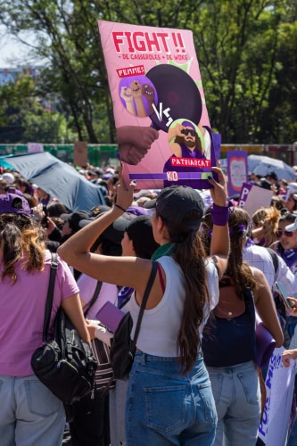 Une manifestation avec des personnes tenant des pancartes, y compris une qui dit 'FIGHT!!' avec des illustrations de femmes se battant contre le patriarcat.