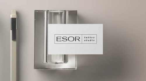Carte de visite blanche avec le texte 'ESOR tattoo studio', placée sur un support transparent, à côté d'un stylo.