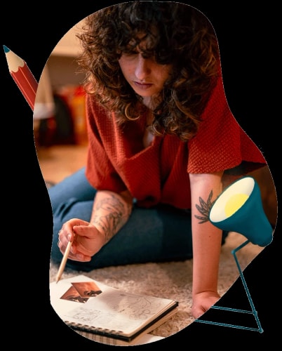 Une femme avec des cheveux bouclés et des tatouages, assise sur le sol avec un carnet de croquis et une lampe à lumière douce, en train de dessiner.