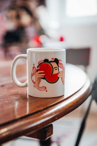 Tasse blanche sur une table en bois, avec un design illustrant une personne tenant des morceaux de pastèque, entourée de gouttes roses.