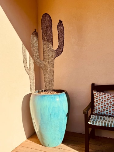 Un grand cactus décoratif en treillis métallique dans un pot en céramique bleu turquoise, placé dans un angle près d'une chaise en bois avec un coussin à motifs, sous une lumière chaude.