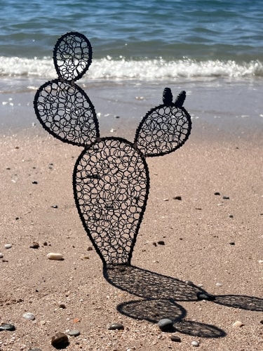 Sculpture en métal représentant un cactus composé de trois segments, posée sur le sable au bord de la mer.