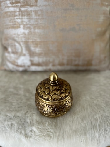 Boîte en métal doré avec un couvercle en forme de bouton, posée sur un coussin en fourrure.