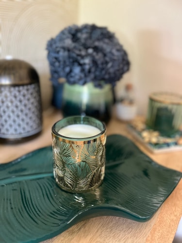 Une bougie parfumée dans un verre décoratif sur un plateau vert, avec un vase de fleurs séchées en arrière-plan et un design intérieur apaisant.
