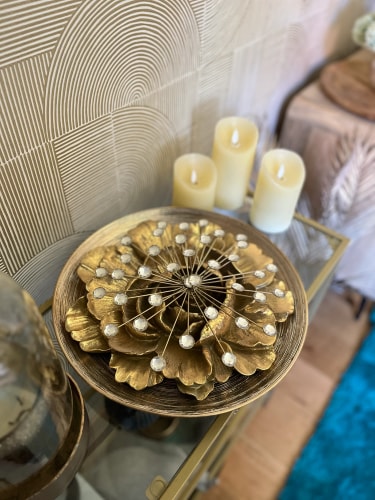 Une assiette décorative en or avec des feuilles et des perles, placée sur une table en verre avec des bougies blanches à côté.