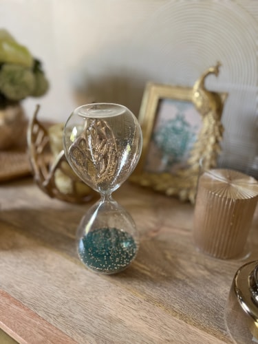 Sablier en verre transparent avec des paillettes turquoise, placé sur une table en bois, entouré de décorations dorées à l'arrière-plan.