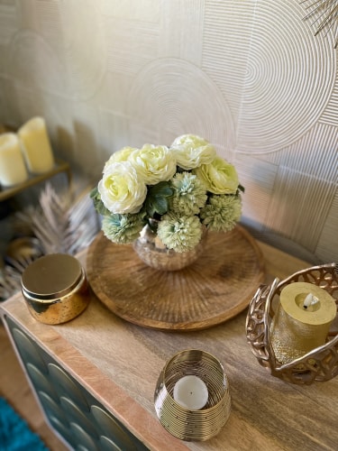Un arrangement floral avec des roses blanches et des fleurs vertes, posé sur une table en bois, entouré de bougies et d'objets décoratifs dorés.