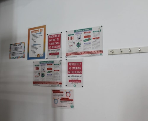 Mur blanc avec plusieurs affiches d’information colorées, principalement en anglais, et une rangée de crochets métalliques fixée à droite.