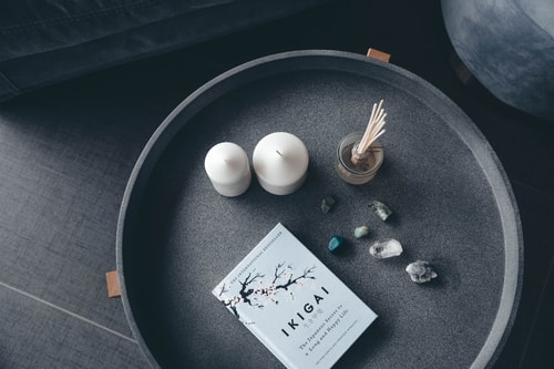 Une table ronde en noir contenant un livre intitulé 'IKIGAI', deux bougies blanches, un vase avec des bâtonnets parfumés et plusieurs pierres colorées.