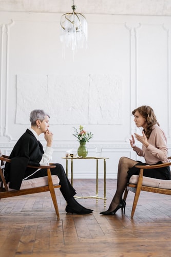 Deux femmes assises dans des fauteuils en bois, en train de discuter dans un intérieur lumineux avec un vase de fleurs sur une table basse.