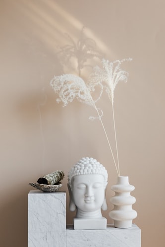 Sculpture de Bouddha blanche posée sur un support en marbre, accompagnée d'un vase en céramique et de branches sèches blanches, avec de la fumée en arrière-plan.