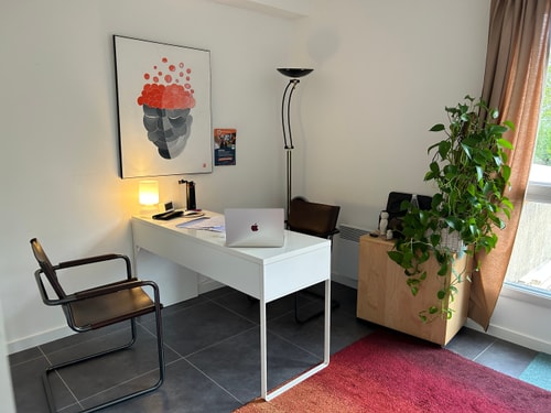 Un bureau moderne dans une pièce au mur clair, avec une chaise, un ordinateur portable sur le bureau, une lampe, et une plante verte dans un coin. Un tableau coloré est accroché au mur.