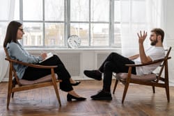 Deux personnes assises dans des fauteuils en bois dans une pièce lumineuse. Une femme écrit sur un bloc-notes tandis qu'un homme parle, gesticulant légèrement. Un horloge est visible sur le mur derrière eux.