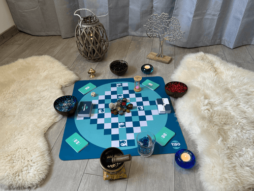 Une vue d'un tapis avec un plateau de jeu du TAO au centre, entouré de pelles avec des pierres colorées, de bougies allumées, et d'une décoration en forme d'arbre. Le tout est posé sur un sol en bois avec des peaux de mouton.