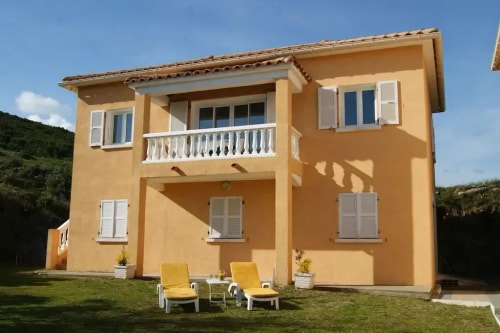 Maison A Pianatella à deux étages avec balcons, escalier et chaises longues devant, sous un ciel bleu.