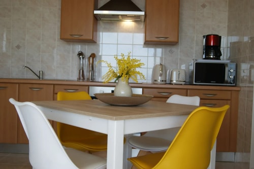 Cuisine moderne avec une table blanche, quatre chaises (deux blanches et deux jaunes), un vase avec des fleurs jaunes, et des appareils électroménagers en arrière-plan.