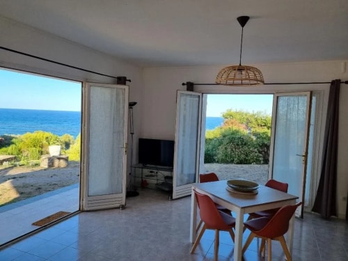 Intérieur d'une maison avec une table et des chaises, vue sur la mer à travers des portes-fenêtres ouvertes, jardin verdoyant visible à l'extérieur.