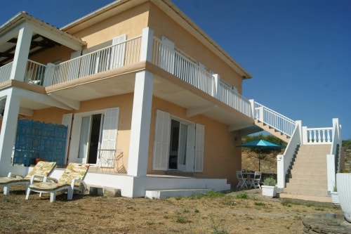 Maison A Pianatella à deux étages avec balcons, escalier et chaises longues devant, sous un ciel bleu.