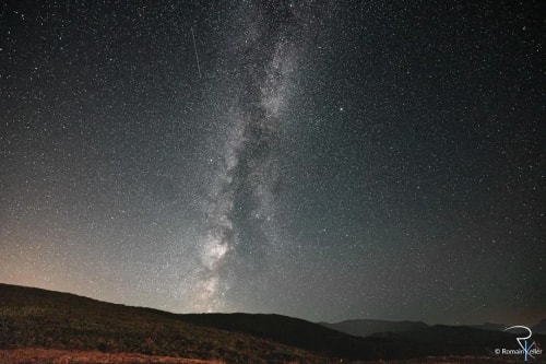 Un ciel nocturne rempli d'étoiles, avec la Voie lactée s'étendant sur le paysage montagneux en bas.