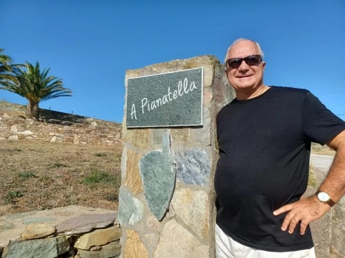 Alain posant à côté d'un panneau indiquant 'A Pianatella', avec des palmiers et un paysage naturel en arrière-plan.