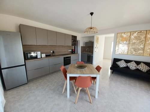 Cuisine moderne avec des armoires grises, un évier et une table à manger blanche entourée de chaises orange. Il y a un réfrigérateur en métal et un canapé noir avec des coussins décoratifs.