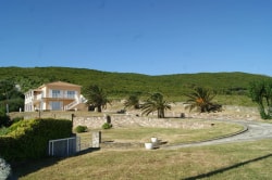 Une maison de style moderne située sur une colline avec des palmiers et un large espace vert, sous un ciel bleu clair.