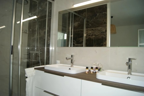 Salle de bain moderne avec deux lavabos, miroir, étagère en bois, et douche en verre.