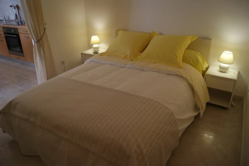 Chambre à coucher avec un lit double aux draps blancs et jaunes, deux lampes de chevet allumées, et un coin cuisine visible en arrière-plan.