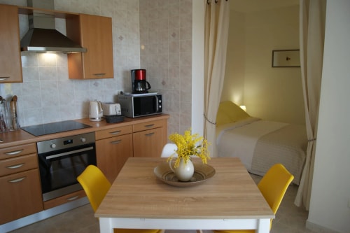 Une cuisine moderne avec des meubles en bois, une table à manger avec des chaises jaunes et un vase de fleurs, et un coin sommeil dans une pièce à côté.