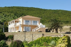 Maison colorée avec un étage située sur une colline verdoyante, entourée de palmiers et de végétation.