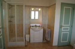 Salle de bains avec douche, lavabo et miroir, murs beige et carrelage, porte verte.