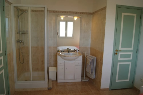 Salle de bains avec douche, lavabo et miroir, murs beige et carrelage, porte verte.
