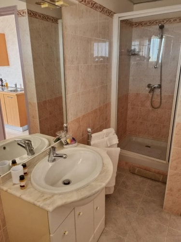 Salle de bain avec deux lavabos en marbre, miroir, douche et serviettes blanches. Le décor est en carrelage beige clair.