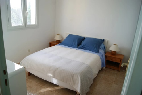 Chambre à coucher avec un lit double et des oreillers bleus, deux lampes de chevet sur des tables de nuit en bois, dans une pièce lumineuse aux murs blancs.