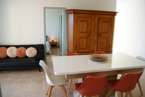 Intérieur d'une cuisine moderne avec un coin salle à manger. La cuisine dispose d'armoires grises, d'un plan de travail en bois, et d'un four. Une table en bois avec des chaises orange est au centre, avec un vase. Un canapé noir avec des coussins décoratifs se trouve à droite. Lumière naturelle entrant par une fenêtre.