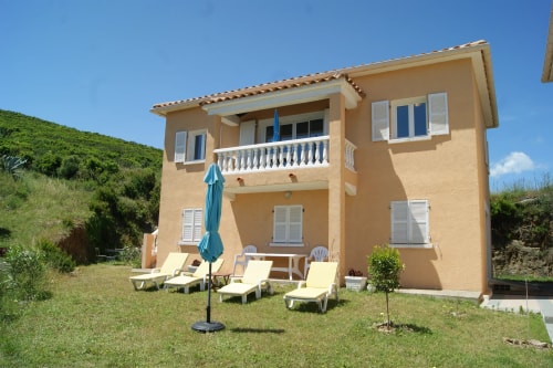 Maison A Pianatella à deux étages avec balcons, escalier et chaises longues devant, sous un ciel bleu.