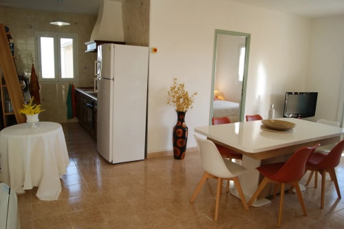 Une cuisine et une salle à manger lumineuses avec une table à manger moderne, des chaises colorées, un réfrigérateur blanc et un coin salon avec un téléviseur.