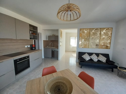 Intérieur d'une cuisine moderne avec un coin salle à manger. La cuisine dispose d'armoires grises, d'un plan de travail en bois, et d'un four. Une table en bois avec des chaises orange est au centre, avec un vase. Un canapé noir avec des coussins décoratifs se trouve à droite. Lumière naturelle entrant par une fenêtre.