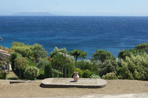 Vue d'un paysage côtier avec un pot en terre cuite sur une plateforme en pierre, entouré de verdure et de la mer bleue en arrière-plan, sous un ciel ensoleillé.