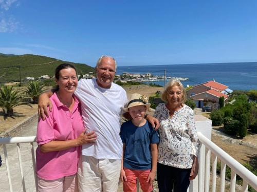 Une famille pose pour une photo sur une terrasse avec vue sur la mer, entourées de paysages verdoyants et de maisons. Il fait beau et ensoleillé.